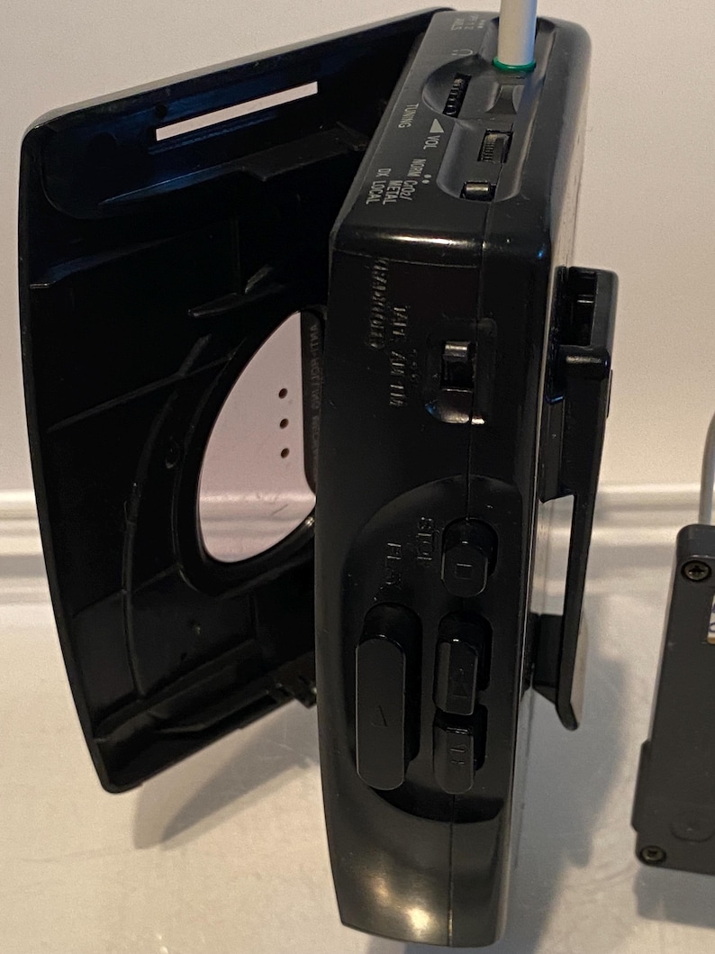 May include: Black portable cassette player with an open lid. The device has buttons for play, stop, and rewind. A white and green audio jack is inserted. The device has a clip on the back.