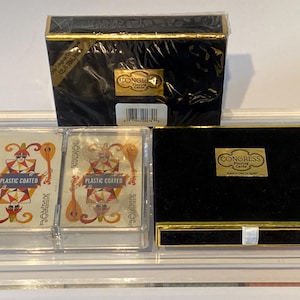 May include: Two decks of Congress Playing Cards in a clear plastic case. The cards are plastic coated and feature a joker card with a colorful jester design. The case is on a black surface with a black box with gold trim that says "Congress Playing Cards" on it.