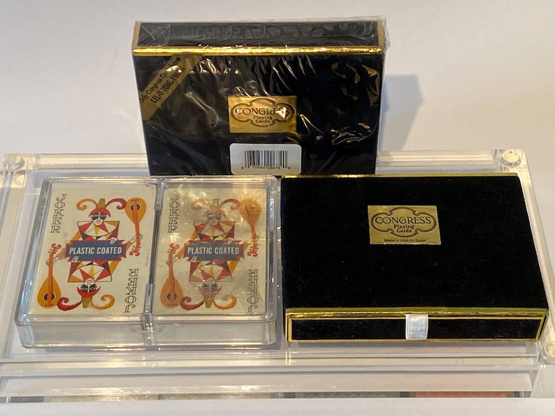 May include: Two decks of Congress Playing Cards in a clear plastic case. The cards are plastic coated and feature a joker card with a colorful jester design. The case is on a black surface with a black box with gold trim that says "Congress Playing Cards" on it.