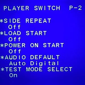 May include: A blue screen displays white text, including "PLAYER SWITCH P-2" and several menu options. The options include "SIDE REPEAT," "LOAD START," "POWER ON START," "AUDIO DEFAULT," and "TEST MODE SELECT."
