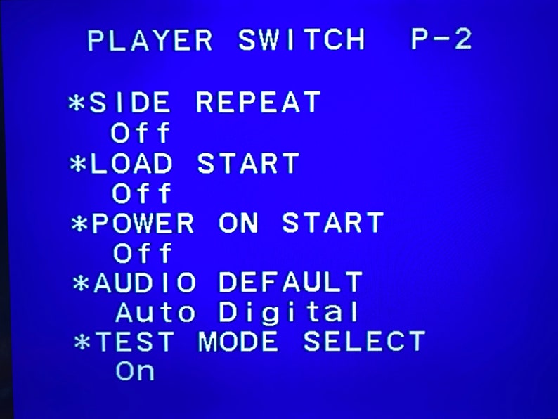 May include: A blue screen displays white text, including "PLAYER SWITCH P-2" and several menu options. The options include "SIDE REPEAT," "LOAD START," "POWER ON START," "AUDIO DEFAULT," and "TEST MODE SELECT."