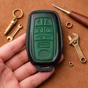May include: A green and black leather car key fob cover with green stitching. The fob has buttons labeled "HOLD" and a key ring and small wrench keychain. A small adjustable wrench and other tools are also visible.