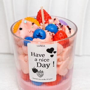 Op de afbeelding: Decoratieve kaars in een helder glas met een roze basis. De kaars is bedekt met roze, oranje, rode en blauwe wasvormen die op fruit lijken. Een label leest "Have a nice Day!" met een bessenillustratie.