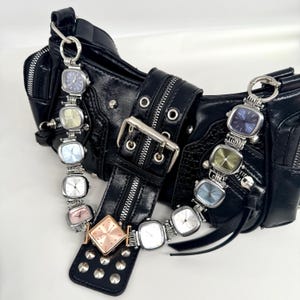 May include: Black leather handbag with silver hardware and multiple watch-style charms. The charms feature square watch faces in various colors, including blue, green, and pink. The bag has a buckle closure and studded accents.