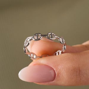 May include: A silver-coloured ring featuring a continuous chain-link design. The band is composed of small, interconnected oval-shaped links, each with a central horizontal bar, creating a uniform, repeating pattern around the entire circumference.