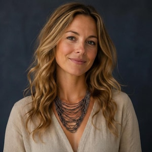 May include: A multi-strand necklace featuring numerous thin, dark metallic chains that drape in a layered, cascading fashion around the neck. The necklace is worn over a plain, light beige V-neck top against a dark, solid blue background.