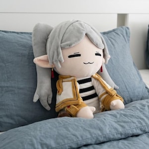 Puede incluir: Un muñeco de peluche con cabello gris claro en coleta, orejas puntiagudas con pequeños pendientes de borla roja y una expresión sonriente. El muñeco viste una camisa a rayas blancas y negras bajo una chaqueta dorada y blanca con hombreras.