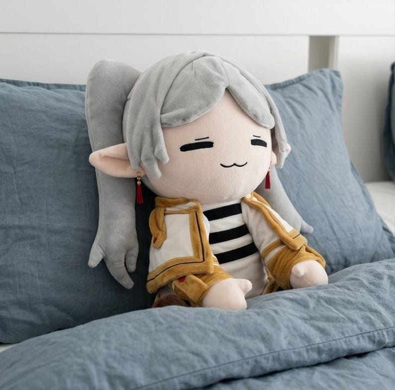 May include: A plush doll featuring light gray hair styled in a ponytail, pointed ears with small red tassel earrings, and a smiling expression. The doll wears a white and black striped shirt under a gold and white jacket with structured shoulders.