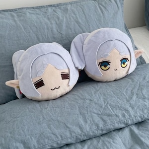 Puede incluir: Dos almohadas de felpa con cabello azul claro y orejas de elfo descansan sobre una cama azul. Las almohadas tienen rasgos faciales bordados, incluidos ojos, cejas y una boca pequeña. Las almohadas están sobre una colcha azul.