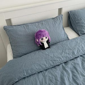 May include: A plush doll with purple hair and a black jacket rests on a blue-grey pillow. The doll has large eyes and a white shirt. The pillow is on a bed with a matching duvet cover. The bed frame is white.