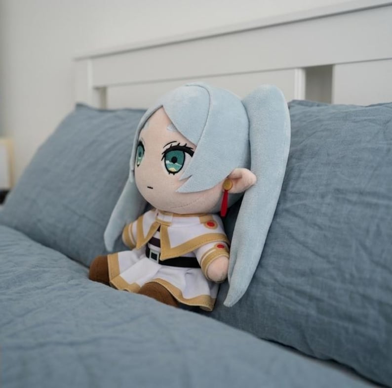 May include: A plush doll featuring light blue hair styled in twin tails, large green eyes, and a neutral expression. The doll wears a white and gold outfit with a black belt, brown shoes, and a single red earring dangling from one pointed ear.