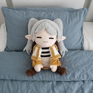 May include: A plush doll featuring a character with light gray hair styled in twin tails, pointed ears, and a neutral expression. The doll wears a white and yellow jacket over a black and white striped shirt, paired with brown boots.