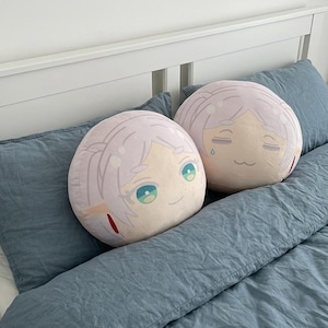 May include: Two round plush pillows with anime-style character faces. The pillows feature light pink hair, teal eyes, and smiling expressions. They rest on a blue bed with a white headboard.