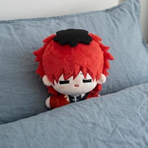 May include: A plush doll with red hair and a black headpiece, resting on a blue pillow. The doll has a sad expression with a tear. The doll is wearing a red outfit with black accents.