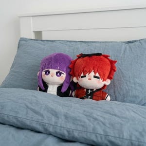 May include: Two plush dolls with anime-style features rest on a blue bed. One doll has purple hair and wears a black jacket over a white shirt. The other doll has red hair and wears a red jacket with black accents.