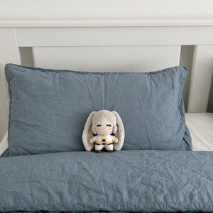 May include: A small plush doll with light gray hair, long floppy ears, and a white and yellow outfit featuring black accents. The doll has a simple embroidered face with closed eyes and a small mouth, positioned on a blue textured pillow.