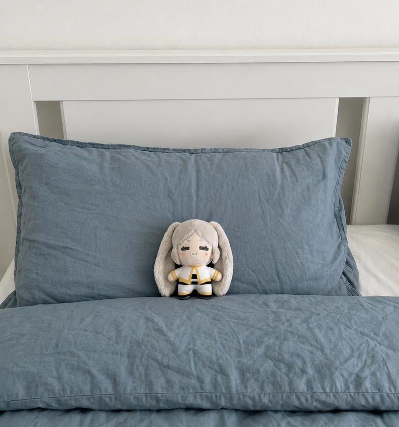 May include: A small plush doll with light gray hair, long floppy ears, and a white and yellow outfit featuring black accents. The doll has a simple embroidered face with closed eyes and a small mouth, positioned on a blue textured pillow.