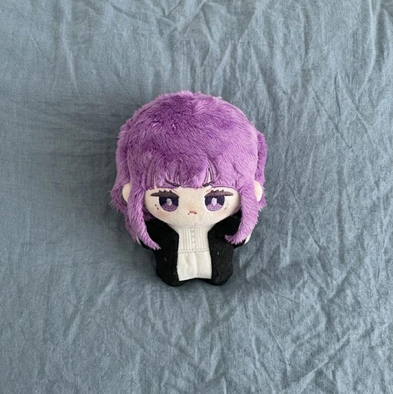 May include: A plush doll head with purple hair and a black jacket over a white shirt. The doll has large eyes and is laying on a blue surface.