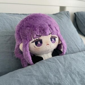 May include: A plush doll with a purple wig and embroidered facial features, including large purple eyes and a small mouth. The doll is resting on a blue pillow, with a white sweater visible at the neck.