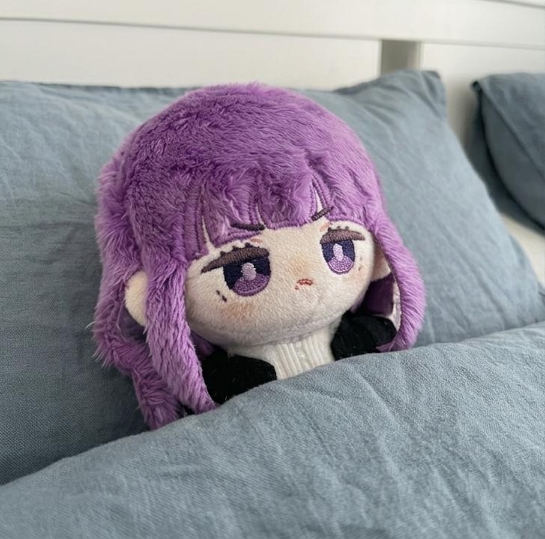 May include: A plush doll with a purple wig and embroidered facial features, including large purple eyes and a small mouth. The doll is resting on a blue pillow, with a white sweater visible at the neck.