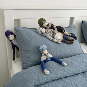 May include: Several anime-style plush dolls rest on a blue bed. One doll with purple hair peeks from the side. Two dolls are on a pillow, one with green hair and black clothing, the other with white hair and a white dress. A blue-haired doll sits on the bed.