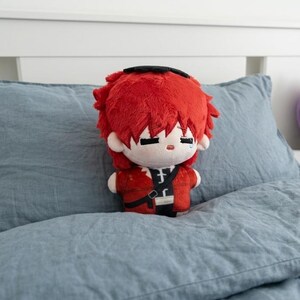 May include: A plush doll with red hair and a red jacket, black pants, and a black strap. The doll has a sad expression with a single tear. It is sitting on a blue pillow.