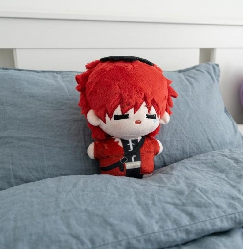 May include: A plush doll with red hair and a red jacket, black pants, and a black strap. The doll has a sad expression with a single tear. It is sitting on a blue pillow.