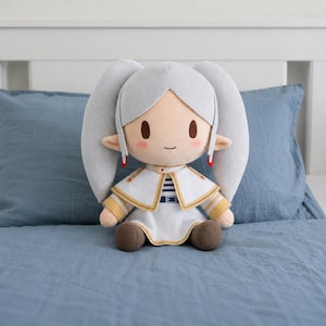 May include: A plush doll featuring a character with long silver hair styled in twin tails, pointed ears, and pink cheeks. The doll wears a white capelet with gold trim over a white dress with blue stripes, a blue belt, and brown shoes.