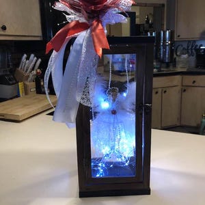 Puede incluir: Una linterna de madera oscura con frente de vidrio, decorada con una rosa roja, encaje blanco y cintas. En el interior, una pequeña figura de ángel está iluminada por luces de hadas azules, creando una exhibición decorativa y festiva.