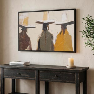 Op de afbeelding: Abstracte kunstprint in een zwarte lijst. Het kunstwerk toont drie figuren met hoeden en jassen in tinten bruin, groen en geel tegen een neutrale achtergrond. Onder het kunstwerk staat een zwarte houten console met twee lades en een onderste plank.