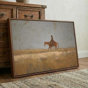 May include: A framed painting depicts a lone cowboy on horseback traversing a vast, muted landscape. The artwork features a palette of earthy tones, with a focus on the rider and horse against a backdrop of a hazy sky. The painting is encased in a dark wooden frame.