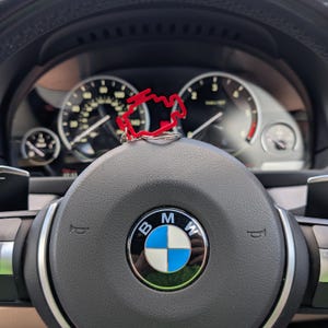 May include: A red engine-shaped keyring sits on a BMW steering wheel. The steering wheel is black with the BMW logo in the centre. The background shows the car's dashboard with gauges and dials.