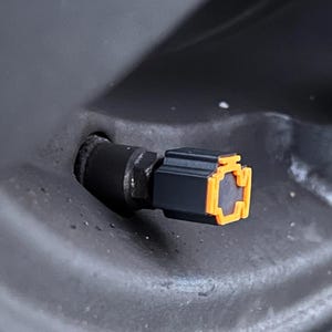 Puede incluir: Un tapón de válvula de neumático negro con perfil cuadrado y un anillo de acento naranja brillante en el borde exterior. El tapón está unido a un vástago de válvula de goma negra que sobresale de una llanta gris oscuro, un componente automotriz.