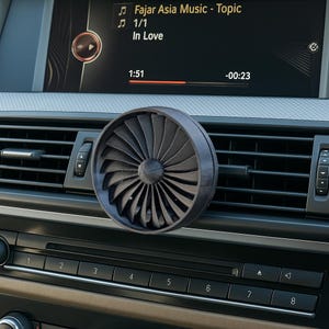 May include: A black turbine-shaped car air vent diffuser. The diffuser is attached to the car's air vent. The car's infotainment screen displays music information. The screen shows the song "In Love" by Fajar Asia Music.