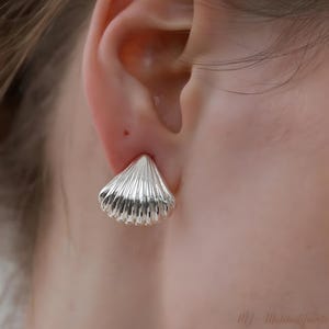 May include: A close-up of a silver seashell-shaped stud earring. The earring is worn on an ear and has a ridged texture. The earring is a bright, shiny silver colour. The earring is a stud style.