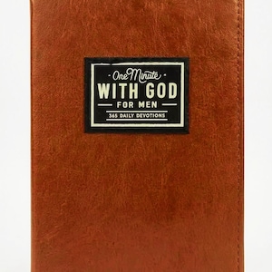 May include: A rectangular book with a textured brown cover. Centered on the front is a black rectangular label with white text that reads: One Minute WITH GOD FOR MEN 365 DAILY DEVOTIONS. The book has stitched edges along the perimeter.