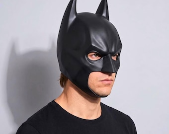 May include: A matte black moulded superhero mask featuring two pointed ears at the top, a contoured brow, and an open face design that exposes the eyes, nose, and mouth area. The mask is constructed from a smooth, rigid material with defined facial features.