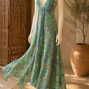 Puede incluir: Un vestido largo tipo halter con un vibrante estampado de cachemira en tonos verde, azul y dorado. La prenda presenta un escote en V profundo, tirantes finos y un dobladillo asimétrico en punta que fluye desde el corpiño ajustado.