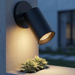May include: A black outdoor spotlight fixture mounted on a concrete wall. The cylindrical light is angled downward, illuminating a small planter with green plants. The fixture has a circular base and a focused beam.