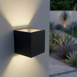 May include: A contemporary black cube-shaped outdoor wall light. The light is mounted on a light grey textured wall, casting a warm glow. The fixture's open top allows light to shine upwards, creating a dramatic effect.