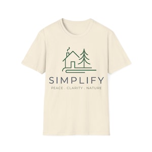 Puede incluir: Camiseta color crema con un gráfico verde y gris. El gráfico presenta un diseño de casa y árbol con la palabra "SIMPLIFY" y las palabras "PEACE . CLARITY . NATURE". La camiseta está hecha de un material suave.
