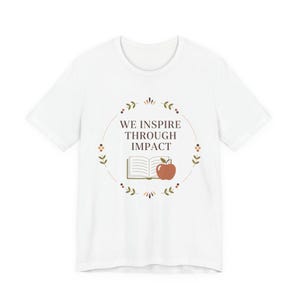 May include: White t-shirt with a round graphic featuring the text "WE INSPIRE THROUGH IMPACT". The graphic includes an open book, a red apple, and decorative floral elements. A casual, everyday tee.