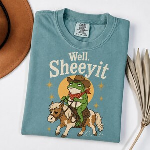 May include: A blue-green short-sleeve t-shirt featuring a graphic of a green frog wearing a brown cowboy hat and red bandana, riding a brown and white pony. Above the graphic, the text reads "Well. SHEEYIT" in a cream-colored font.
