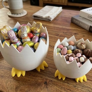 Peut inclure: Deux bols blancs en forme d'œuf avec des pieds jaunes, remplis de friandises de Pâques. L'un contient des lapins et des œufs en chocolat emballés, l'autre des bonbons et chocolats mouchetés. Les bols sont sur une table en bois.