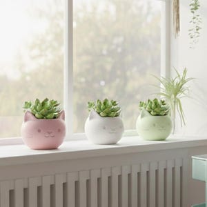 May include: Three cat-shaped ceramic planters in pink, white, and green, each holding a succulent plant. The planters sit on a white windowsill, with a window and greenery in the background.