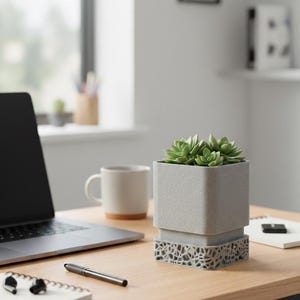 Pode incluir: Um vaso quadrado cinza claro com uma base geométrica, contendo suculentas verdes. O vaso está em uma mesa de madeira com um laptop, caneta, bloco de notas e caneca.