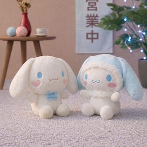 May include: Two plush toys, one white and one white with a light blue hood, sit on a light-colored carpet. The white toy has a blue bow tie and pink heart cheeks. The other has blue ears and a matching hood. A sign with Japanese text hangs in the background.