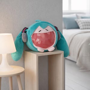 May include: A teal plush toy with a large open mouth design, resembling a cartoon character. The toy has long teal pigtails, gray headphones, and a pink interior mouth. It sits on a light wooden shelf, with a lamp and bed in the background.