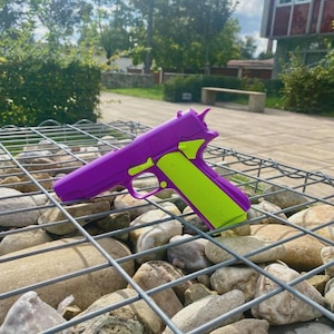 Puede incluir: Una pistola de juguete morada con detalles en verde neón. La pistola de juguete está sobre una estructura de malla metálica llena de piedras. El fondo muestra vegetación y un edificio.