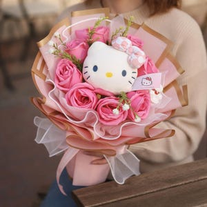 Könnte beinhalten: Ein Strauß aus rosa Rosen und Grünzeug, verpackt in rosa und goldenem Papier, gebunden mit einem rosa Band. Ein weißes Hello Kitty-Plüschtier mit einer rosa Schleife ist zwischen den Blumen eingebettet. Ein kleines Etikett mit dem Hello Kitty-Logo ist angebracht.